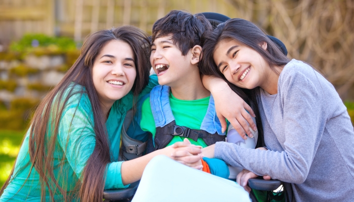 Sisters laughing and hugging disabled little nine year old brother in wheelchair outdoors.