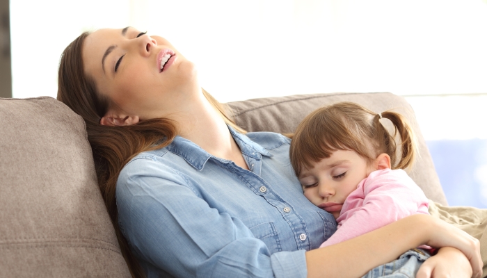 Tired mother sleeping embracing to her asleep baby daughter sitting on a sofa at home.