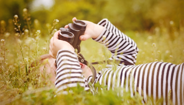 Young child using a pair of binoculars to look up into the sky as he lies on back in a grassy rural meadow enjoying a day in nature.