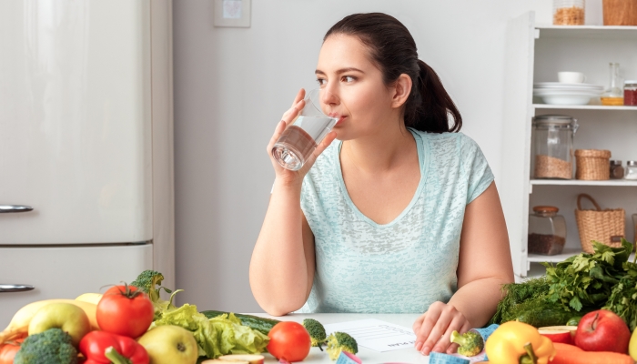 Young chubby woman standing in kitchen sitting at table holding glass drinking pure water looking out the window pensive.