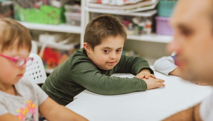 Young cute boy with down syndrome in green shirt sitting at white desk with other kids and studying.