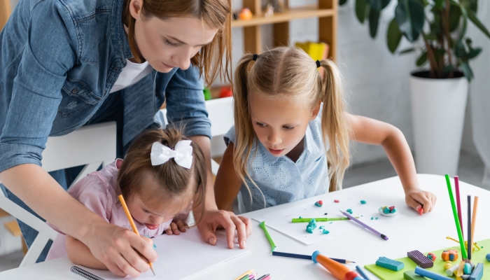 Teacher assisting disabled child with down syndrome drawing in private kindergarten.