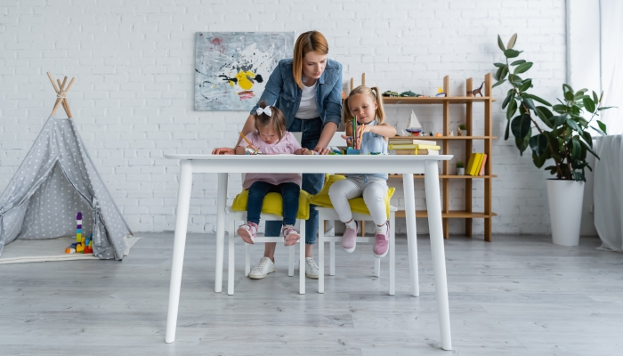 Teacher standing behind preschooler girl and disabled child with down syndrome drawing in private kindergarten.