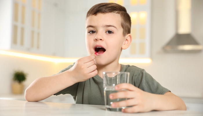 Little boy with glass of water taking vitamin capsule in kitchen.
