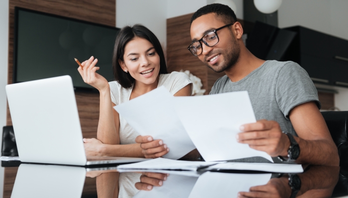 Photo of cheerful loving young couple using laptop and analyzing their finances with documents.