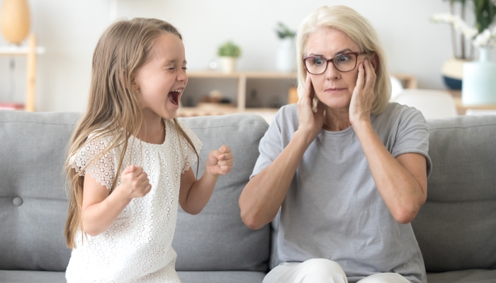 Shocked grandma closing ears not to hear noisy stubborn fussy little granddaughter.