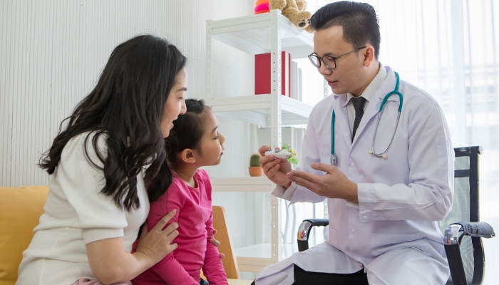 Young Asian man in glasses with stethoscope explain about medical equipment to little girl sitting with mother in office.