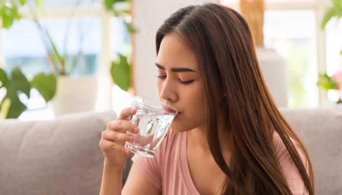 Young Asian woman drinking water and taking pill while sitting on sofa at home.
