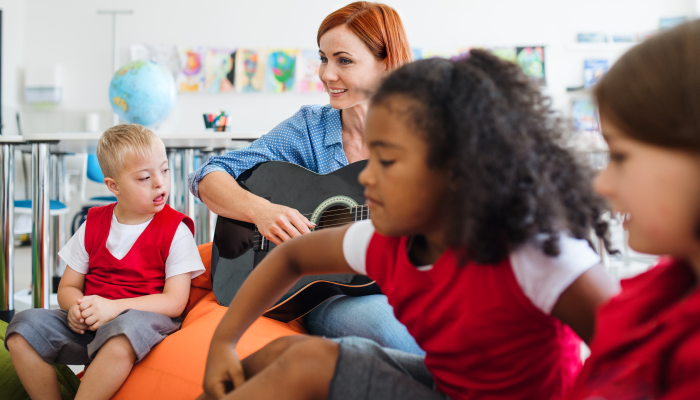 A group of small school kids and teacher with guitar sitting on the floor in class.