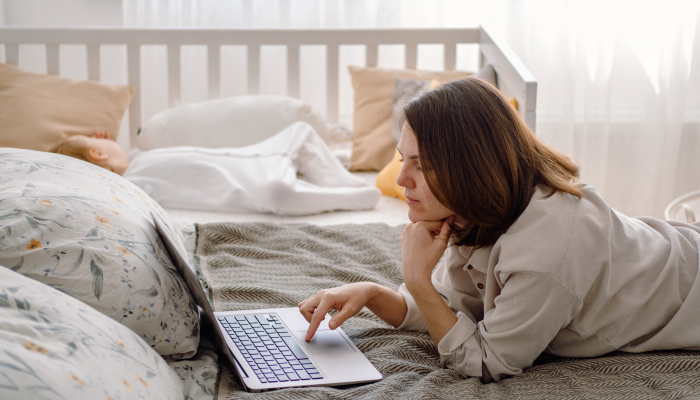 A young mother works on laptop during quiet time while her baby toddler sleeps on the crib.