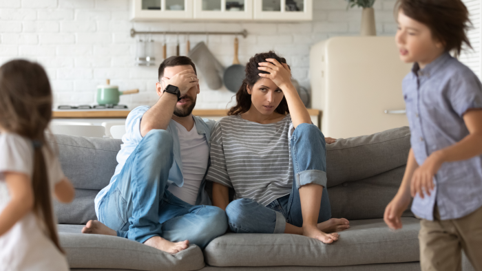 Annoyed young parents sit on couch in kitchen tired from loud two little children running playing.