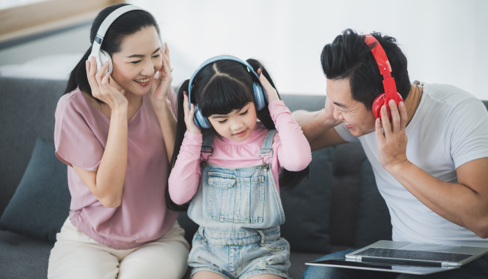 Asian parent mom and dad with female chid are listening to music with wireless headphones and laptop at home.