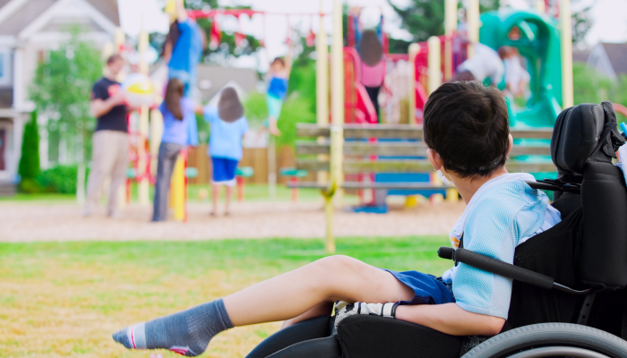 Disabled little boy in wheelchair sadly watching children play on playground.