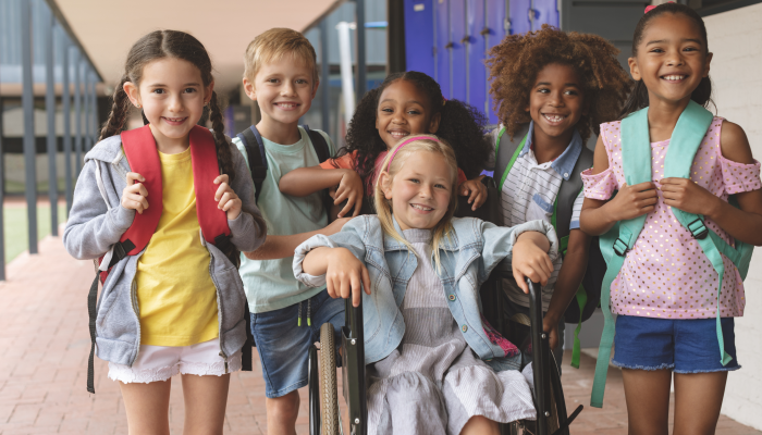 Front view of happy diverse school kids standing in outside corridor at school while a Caucasian schoolgirl is sitting on wheelchair in foreground.