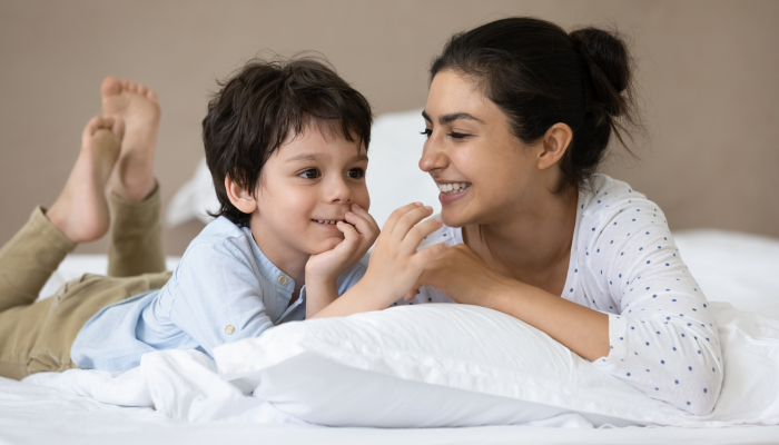 Happy Indian little boy and cheerful mom resting in bed, talking, laughing, having fun, enjoying leisure together in weekend morning.