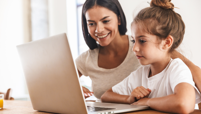 Image of attractive family woman and her little daughter smiling and using laptop computer together while sitting at table in apartment.