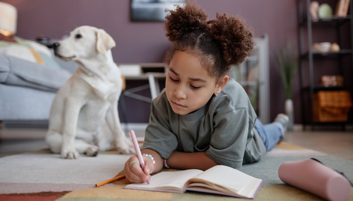 Portrait of black preteen girl writing in diary lying on floor at home with dog in background.