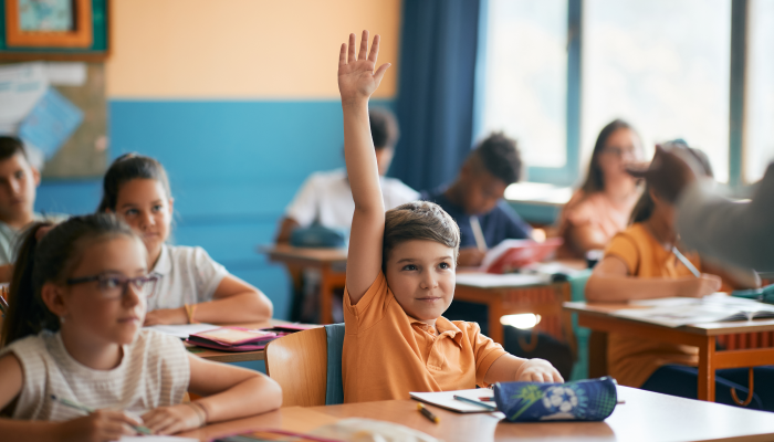 Smiling elementary student raising his hand to answer a question during class at school.