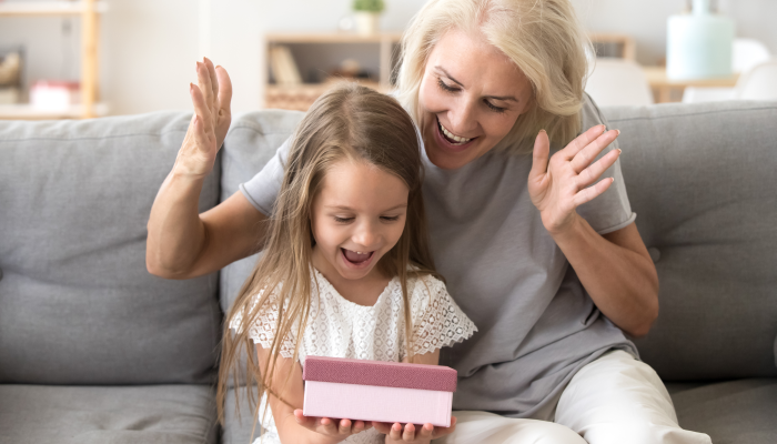 Smiling grandmother making surprise to cute little granddaughter giving gift box.