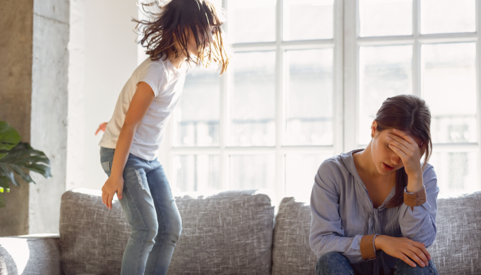 Upset mother having problem with noisy naughty daughter jumping on couch and screaming.