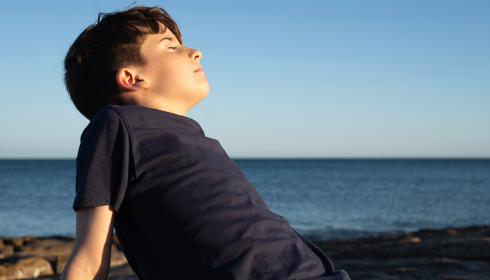 Young kid enjoying good weather on the coast, shore, breathing deep fresh sea air with eyes closed during sunset.