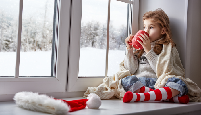 Little girl sitting by the window with a cup of hot drink and looking at the winter forest.