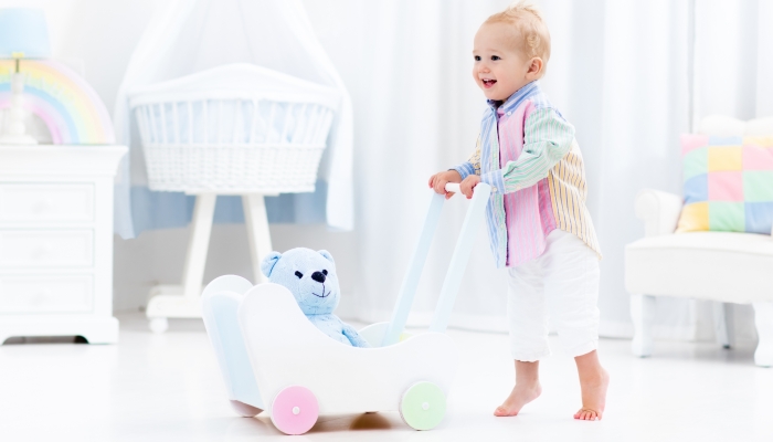 Baby boy learning to walk with wooden push walker in white bedroom with pastel rainbow color toys.