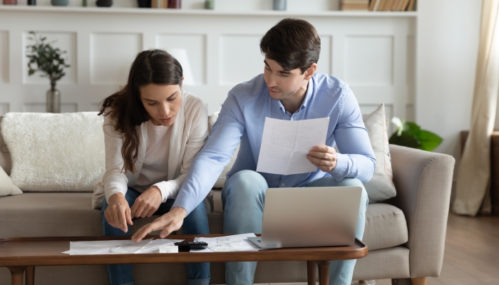 Concentrated attentive millennial spouses sitting on couch at home discussing possibility of taking out loan or credit.