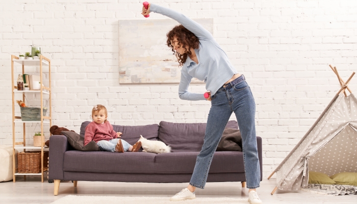 Curly woman exercising with dumbbells near toddler daughter on couch in cozy living room.