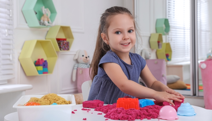 Cute little girl playing with bright kinetic sand at table in room.