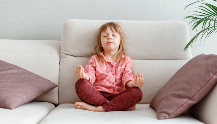 Little caucasian 3 year girl meditates on sofa while practicing yoga.