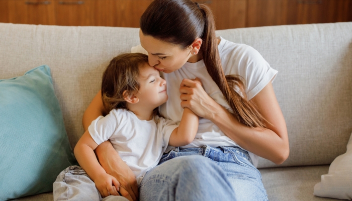 Loving Mom Kissing And Embracing Her Little Daughter While Sitting Together On Couch.