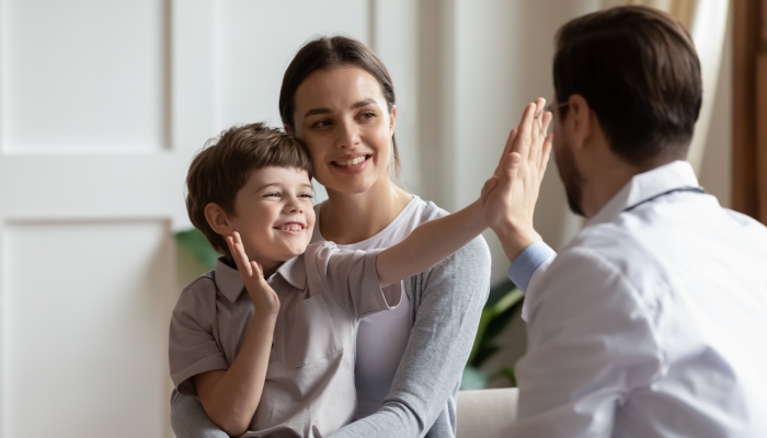 Overjoyed little boy patient have fun give high five to male doctor at consultation in clinic with mom.