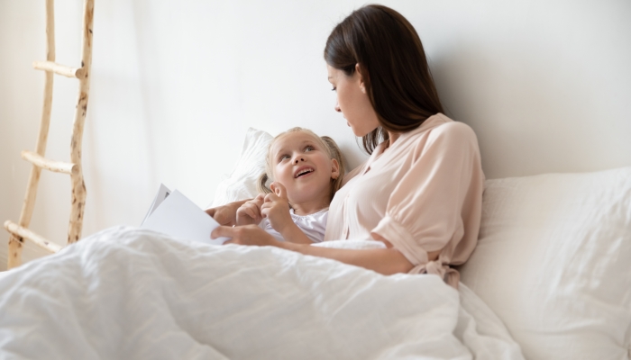 Overjoyed little preschool child girl lying under blanket with young mommy, asking questions about story heroes.