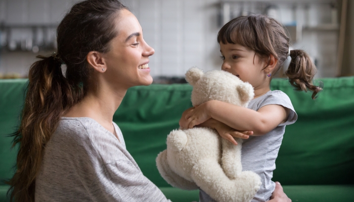 Smiling mother hugging cute little girl holding teddy bear toy showing love and care in family.