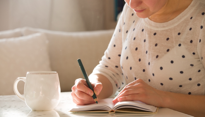 Woman sitting at the table, writing in the notebook and drinking coffee in nice light home interior.