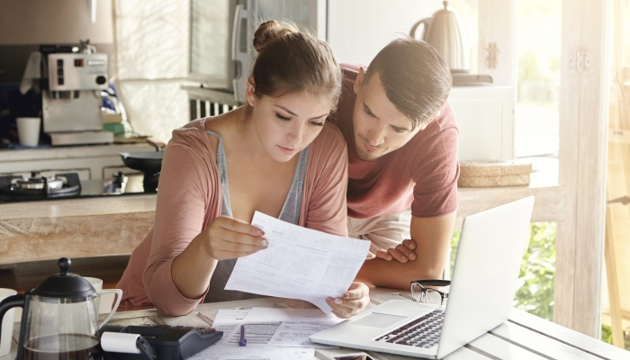 Young couple managing finances, reviewing their bank accounts using laptop computer and calculator at modern kitchen.