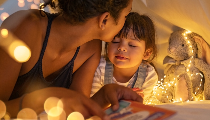 African american mother kissing her daughter on forehead while lying on bed in illuminated tent.