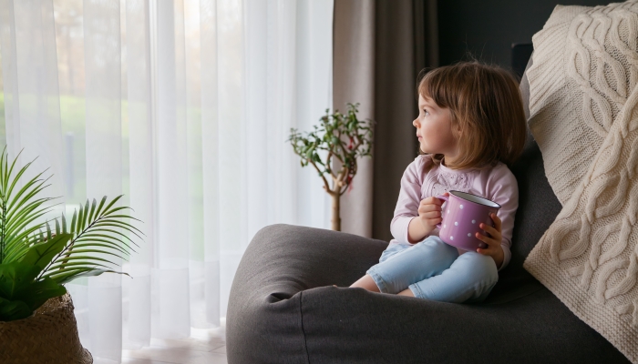 Little cute baby girl with a cup of tea looking through the window sitting on a cozy bean bag chair.